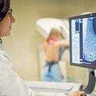 A female doctor examines scans from a mammogram. In the background is a woman standing at the mammogram machine.