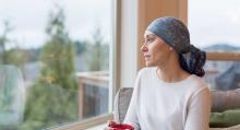 A young woman with cancer sits by her living room window and gazes out contemplatively. She is wearing a headscarf and drinking a cup of tea.
