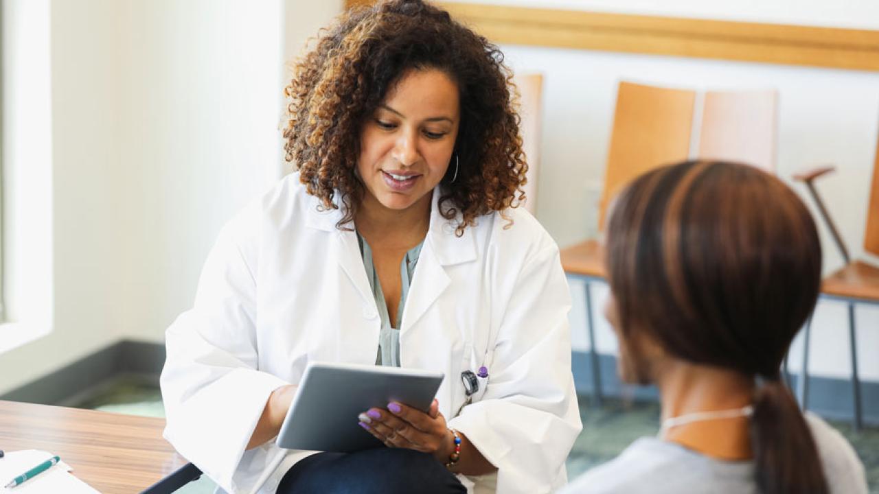 A Black female doctor holds a digital tablet as she consults with a female patient.