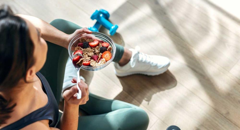 A woman in athletic gear sits while eating a bowl of fruit and oats.