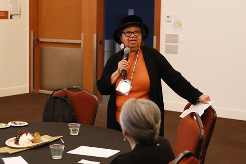Woman holding a microphone standing at a round table while addressing the room