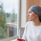 A young woman with cancer sits by her living room window and gazes out contemplatively. She is wearing a headscarf and drinking a cup of tea.